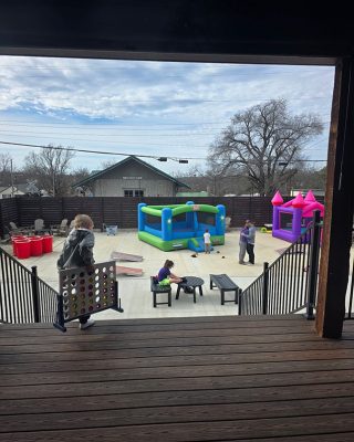 A woman stands on a wooden deck, gazing at a playground filled with children playing.